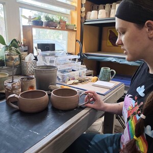 May include: Two unfinished, light brown ceramic mugs with rounded bodies and attached handles sit on a work surface. The mugs are in a pottery studio, with shelves of finished mugs in the background. A person is holding one of the mugs.