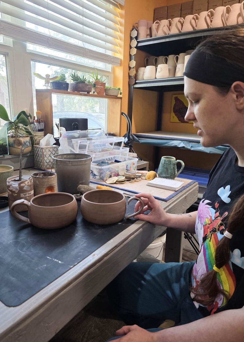 May include: Two unfinished, light brown ceramic mugs with rounded bodies and attached handles sit on a work surface. The mugs are in a pottery studio, with shelves of finished mugs in the background. A person is holding one of the mugs.