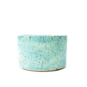 May include: A cylindrical ceramic bowl featuring a light turquoise glaze with a speckled pattern of dark brown and black dots. The vessel has a smooth, straight-sided exterior and a flat base, showcasing a consistent color gradient throughout.