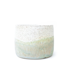 May include: A cylindrical ceramic planter featuring a speckled white upper section and a smooth, light green glazed lower section. The vessel has a straight-sided profile with a textured finish, showcasing a distinct color transition between the top and bottom halves of the exterior surface.