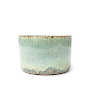 May include: A cylindrical ceramic bowl featuring a reactive glaze with a gradient of soft mint green and pale sage tones. The upper rim displays a darker, speckled brown drip pattern, while the base shows subtle blue-grey textured accents.
