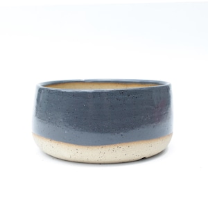 May include: A handcrafted ceramic bowl with a two-tone design. The upper portion is a matte navy blue, while the lower section is a speckled beige. The bowl has a rounded shape and a smooth, glazed finish, perfect for serving or display.