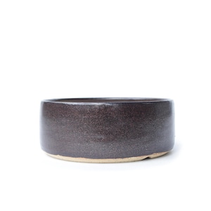 May include: A handcrafted, dark brown ceramic bowl with a textured surface. The bowl has a cylindrical shape with a slightly lighter-colored base. This piece is suitable for serving or as a decorative accent.