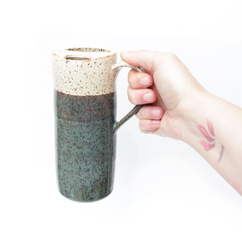 Puede incluir: Una taza de cer&aacute;mica alta con un esmalte blanco y marr&oacute;n moteado en la mitad superior y un esmalte verde oscuro en la mitad inferior. La taza tiene un asa y un peque&ntilde;o agujero en la parte superior.