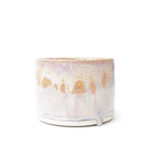 Puede incluir: Una maceta cilíndrica de cerámica con un esmalte reactivo que combina tonos lavanda suave, rosa pálido y blanco crema. La superficie muestra una textura variada y goteada con motas de arcilla color canela visibles en la base.