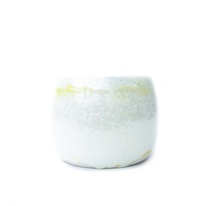 May include: A small, round ceramic cup with a speckled, off-white glaze. The rim has a subtle yellow accent. The cup's shape is slightly wider at the base, tapering towards the top. The overall aesthetic is minimalist and handcrafted.