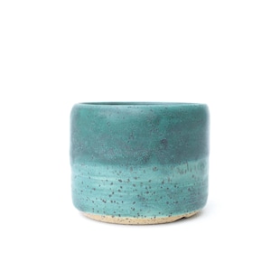 May include: A handcrafted, cylindrical ceramic pot with a teal and turquoise glaze. The pot has a speckled texture and a natural, unglazed base. The design is simple and modern, suitable for plants or decor.