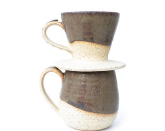 May include: A ceramic coffee pour-over dripper stacked on top of a matching ceramic mug. Both pieces feature a two-tone design with a dark brown, speckled upper section and a cream-colored, speckled lower section, each with a curved handle.