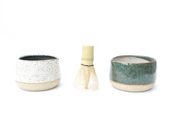 May include: A matcha tea set featuring a bamboo whisk, a speckled white ceramic bowl, and a teal ceramic bowl. The bowls have a natural, unglazed base. The whisk is made of light-colored bamboo.