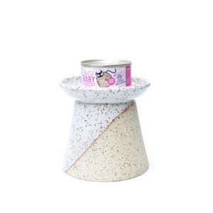 May include: A speckled ceramic cat food bowl with a pedestal base. The base is divided diagonally, with a white and beige color scheme. A can of cat food with pink and silver labels sits on top of the bowl.