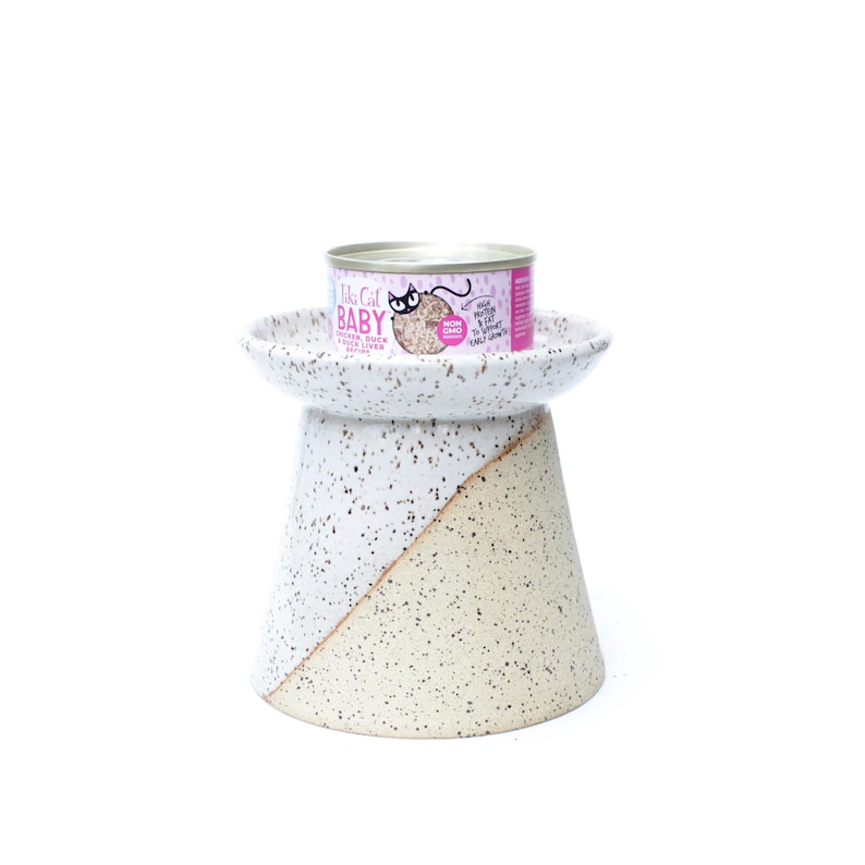 May include: A speckled ceramic cat food bowl with a pedestal base. The base is divided diagonally, with a white and beige color scheme. A can of cat food with pink and silver labels sits on top of the bowl.