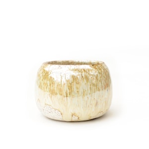 Puede incluir: Un cuenco de cerámica redondo con un esmalte brillante y un patrón de goteo en tonos crema, beige y marrón claro. La parte inferior del cuenco tiene un acabado texturizado y moteado, mientras que la sección superior muestra goteos verticales.