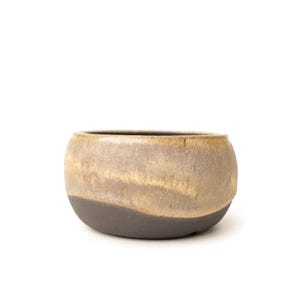 May include: A small, round ceramic bowl featuring a two-tone design. The upper portion has a textured, mottled beige and tan glaze, while the lower section is finished in a solid, dark charcoal gray matte glaze with a visible grainy surface texture.