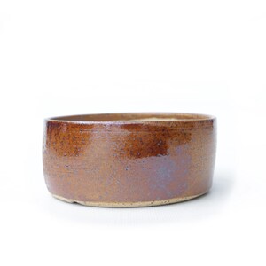 May include: A handmade, brown ceramic bowl with a wide, shallow design. The bowl has a rustic, textured surface with variations in color, ranging from light tan to deep brown. The interior and exterior are glazed, giving it a glossy finish.