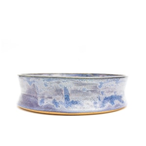 Puede incluir: Un cuenco de cerámica ancho y poco profundo con un esmalte azul claro y un sutil patrón moteado. El cuenco tiene un borde ligeramente acampanado y una base redondeada.