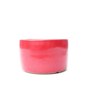 May include: A cylindrical ceramic bowl with a vibrant, glossy red glaze featuring subtle dark speckles throughout the surface. The vessel has a flat base and straight, vertical walls, presenting a simple, functional design for holding items.