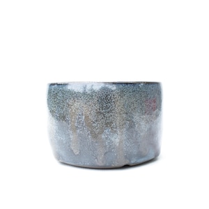 May include: A cylindrical ceramic bowl featuring a reactive glaze with a mottled finish. The exterior displays a blend of deep blue, grey, and earthy brown tones, with a textured, speckled appearance and subtle vertical drips of glaze throughout.