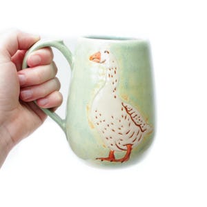 May include: A light green ceramic mug featuring a raised, hand-painted white goose with orange feet and an orange beak. The mug has a curved handle and a smooth, glossy finish, showcasing a detailed illustration of the bird on the front surface.