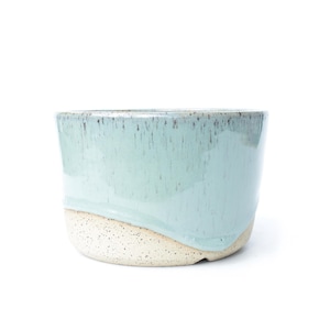 May include: A ceramic planter with a light blue glaze over a speckled, off-white base. The glaze drips down the sides, creating a textured effect. The planter has a rounded shape and a wide opening.