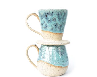 May include: A ceramic coffee pour-over dripper stacked on top of a matching mug. Both pieces feature a speckled cream base with a glossy, mottled turquoise glaze on the upper sections. Each item has a curved handle and a smooth, rounded shape.
