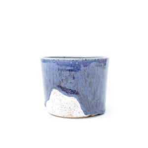May include: A handmade ceramic cup with a unique design. The cup features a dark blue glaze that transitions to a lighter blue and white speckled base. The cup is cylindrical and has a smooth, glossy finish.