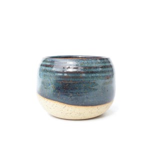 May include: A rounded ceramic cup featuring a two-tone glaze. The upper portion is a deep, glossy blue with subtle dark brown horizontal striations, while the lower section is a speckled, matte off-white with a textured, granular appearance.