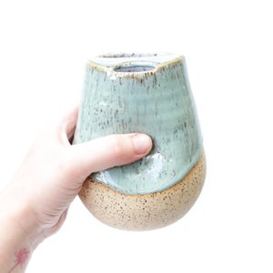 May include: A ceramic cup featuring a two-tone design with a glossy, speckled light green glaze on the upper portion and a textured, speckled beige clay base. The vessel has a rounded, tapered shape with a wide opening at the top.