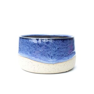 May include: A small, round ceramic bowl with a two-tone design. The top half is a textured, deep blue, while the bottom half is a speckled, off-white. The bowl has a simple, modern aesthetic, suitable for various uses.