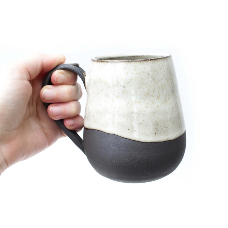 May include: A handmade ceramic mug with a two-tone design. The bottom half is a dark charcoal color, while the top half is a speckled cream color. The mug has a large, curved handle and a smooth, glossy finish.