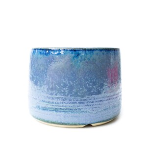 May include: A cylindrical ceramic planter featuring a reactive glaze that transitions from a deep, saturated cobalt blue at the rim to a lighter, speckled periwinkle blue toward the base, with a smooth, glossy finish and a flat, unglazed bottom edge.