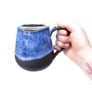 以下が含まれることがあります： ユニークなデザインの手作りセラミックマグ。上部はまだらの青色で、無地の黒色のベースに変わります。マグは丸みを帯びた形状で、黒いハンドルが付いています。マグは手で持たれています。