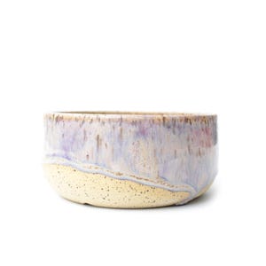 May include: A round ceramic bowl featuring a two-tone glaze. The upper portion displays a smooth, glossy finish in shades of lavender, light blue, and white, while the lower section has a textured, speckled cream-colored matte surface.