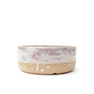 Puede incluir: Un cuenco de cerámica de perfil bajo con una base beige moteada y un esmalte texturizado y goteante en tonos blanco y malva apagado. La superficie exterior muestra un contraste marcado entre la base de arcilla cruda y la parte superior brillante.