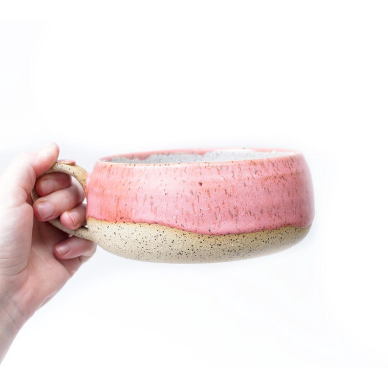 May include: A handmade ceramic mug with a wide, shallow bowl and a large handle. The mug features a speckled, light beige base and a pink upper half. The interior is a light gray color. The mug is held against a white background.