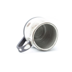 May include: A handmade ceramic mug with a white exterior and a dark gray rim and handle. The interior is gray. The handle features a white dot design. The mug is tilted, showing the inside.