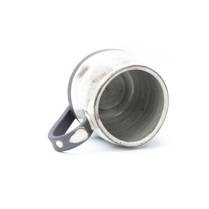 May include: A handmade ceramic mug with a white exterior and a dark gray rim and handle. The interior is gray. The handle features a white dot design. The mug is tilted, showing the inside.