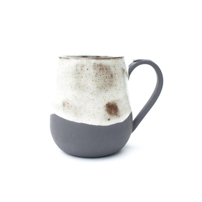 May include: A handmade ceramic mug with a rounded base and a curved handle. The bottom half is a matte gray, while the top half is speckled white and brown. The mug is designed for hot beverages.