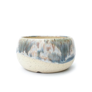 May include: A round ceramic bowl featuring a speckled cream-colored base that transitions into a smooth, glossy glaze with blended streaks of blue, grey, and soft brown tones. The bowl has a wide, open top and a curved, deep basin structure.