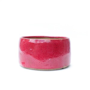 May include: A round, red ceramic bowl with a glossy finish. The bowl has a wide opening and a slightly textured surface. The interior is a lighter, natural clay color. The bowl is suitable for serving or display.