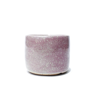 Peut inclure: Une tasse cylindrique en céramique avec un émail moucheté mauve et blanc. La surface extérieure présente une finition texturée et tachetée avec des nuances variées de rose et de blanc, créant un aspect pointillé sur tout le récipient.