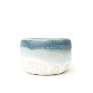 May include: A ceramic bowl featuring a speckled cream-colored base that transitions into a smooth, light blue glaze. The upper rim is a darker blue with visible horizontal ridges, and a white glaze drips down the sides over the speckled texture.