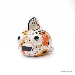 May include: A ceramic fish-shaped container with a lid. The container is painted with orange, yellow, black, and white spots. The fish has large, round eyes and a wide, open mouth.