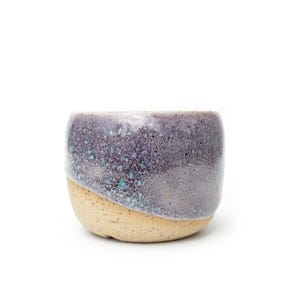 Puede incluir: Una taza de cerámica de forma redondeada, con una base beige moteada y un esmalte texturizado y jaspeado en tonos púrpura oscuro, gris y toques de azul cerceta en la parte superior. La superficie presenta un acabado brillante y variado.