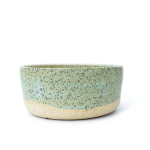 May include: A handmade ceramic bowl with a speckled, light green glaze. The bowl has a rounded shape with a natural, unglazed beige base. The interior and upper portion of the bowl are a speckled green color.