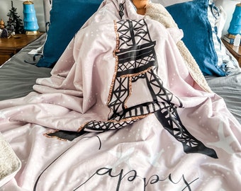Può includere: Una coperta in pile rosa con un designo della Torre Eiffel in bianco e nero e la parola "happy" scritta in corsivo nero. La coperta è su un letto con cuscini blu.