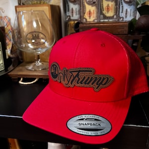 May include: Red and black baseball cap with a brown leather patch that says "On Trump" and a silver sticker that says "The Authentic YUPOONG Snapback YUPOONG Since 1974".