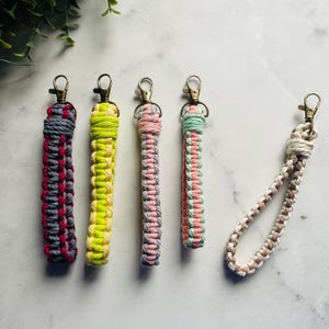 Könnte beinhalten: Eine Sammlung von Makramee-Schlüsselanhängern in verschiedenen Farben, darunter Grau, Rosa, Gelb und Mintgrün. Jeder Schlüsselanhänger hat einen Metallverschluss zur Befestigung. Die Schlüsselanhänger sind auf einer weißen Marmoroberfläche angeordnet.