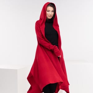 May include: A long, flowing red hooded coat. The coat has a wide, asymmetrical hemline and long sleeves. The model is wearing a black turtleneck and black pants with black boots. The background is white.