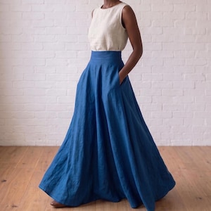 May include: A sleeveless cream-colored top paired with a high-waisted, floor-length blue skirt. The skirt features a wide waistband, a full, flowing silhouette, and side pockets. The ensemble is composed of a solid-colored, textured fabric.