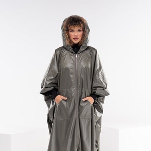 May include: A woman wearing a long, hooded, olive green raincoat with a zipper closure. She is standing on a white surface and wearing black boots.